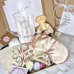 May include: A gift box with items for a "Maid of Honor". The box contains a sleep mask, hair clip, a bag with "Maid of Honor" text, Prosecco gummies, tea, a candle, and a card that says "Will you be my maid of honor?"