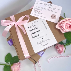 May include: A brown cardboard gift box decorated with a light pink satin ribbon bow. A white card on top reads, I'm here to make your day better, Sealed with love, Lovely things inside! Open me! A second card and two light pink artificial roses.