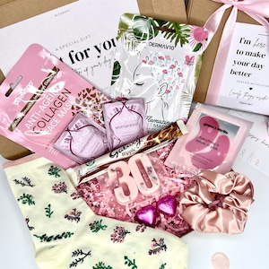 May include: A gift box containing a pair of cream socks with floral patterns, a pink satin scrunchie, two Pukka tea bags, a Galaxy Ripple chocolate bar, a number 30 candle, two heart-shaped chocolates, and three skincare face masks.