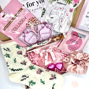 May include: A gift box containing a pair of cream-colored socks with floral patterns, two Pukka tea bags, a Galaxy Ripple chocolate bar, a white number 21 candle, two pink heart-shaped candies, a satin scrunchie, and three skincare face masks.