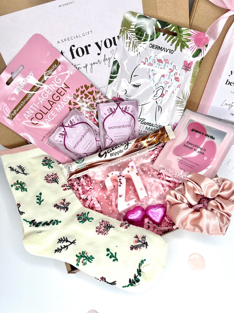 May include: A gift box containing a pair of cream-colored socks with floral patterns, two Pukka tea bags, a Galaxy Ripple chocolate bar, a white number 21 candle, two pink heart-shaped candies, a satin scrunchie, and three skincare face masks.