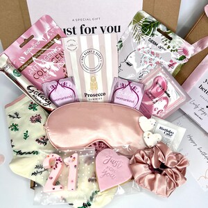May include: A gift box containing a pink satin sleep mask, a matching pink scrunchie, a pair of cream socks with floral patterns, two Pukka tea bags, a Dermavio face mask, Prosecco gummies, a heart-shaped cookie, and a number 21 candle.