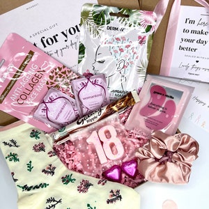 May include: A pink and white gift box filled with various items, including a pair of socks with a floral pattern, a pink face mask, a pink candle, and a pink hair scrunchie. The box is decorated with pink tissue paper and a pink ribbon. The text "I'm here to make your day better" is printed on the box.