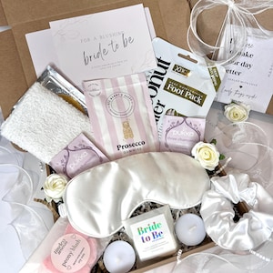 May include: A white gift box filled with various items for a bride-to-be. The box includes a white eye mask, a white satin scrunchie, a pink bath bomb, two white tea lights, a white card with the text "Bride to Be", a pink and white striped bag with gold confetti, a white foot mask, a white tea bag, and a pink and white striped bag with gold confetti.