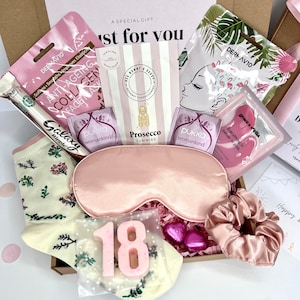 May include: A pink and white gift box filled with various items, including a pink sleep mask, a pair of white socks with floral designs, a pink face mask, a pink hair scrunchie, a pink and white chocolate bar, and a pink number 18 candle.