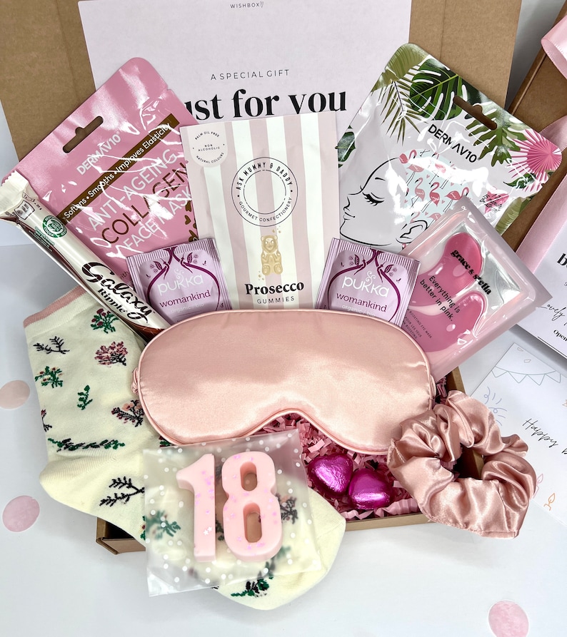 May include: A pink and white gift box filled with various items, including a pink sleep mask, a pair of white socks with floral designs, a pink face mask, a pink hair scrunchie, a pink and white chocolate bar, and a pink number 18 candle.