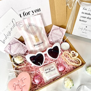 Op de afbeelding: Een cadeauverpakking met een hartvormige zonnebril, een armband, een koekje en Prosecco gummies. De doos bevat een kaart met de tekst "Will You Be My Bridesmaid". De doos is gevuld met roze papier.