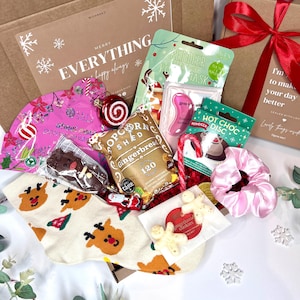 May include: A Christmas-themed gift box filled with festive items. Includes a reindeer face mask, gingerbread popcorn, hot chocolate disc, candy cane, scrunchie, gingerbread cookies, and a pair of reindeer socks. The box is decorated with snowflakes and a red bow.