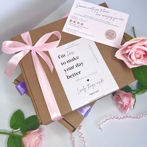 May include: A brown cardboard box with a pink ribbon and a white card that says "I'm here to make your day better." The card is sealed with a heart and says "Lovely things inside! Open me!" The box is surrounded by pink roses.