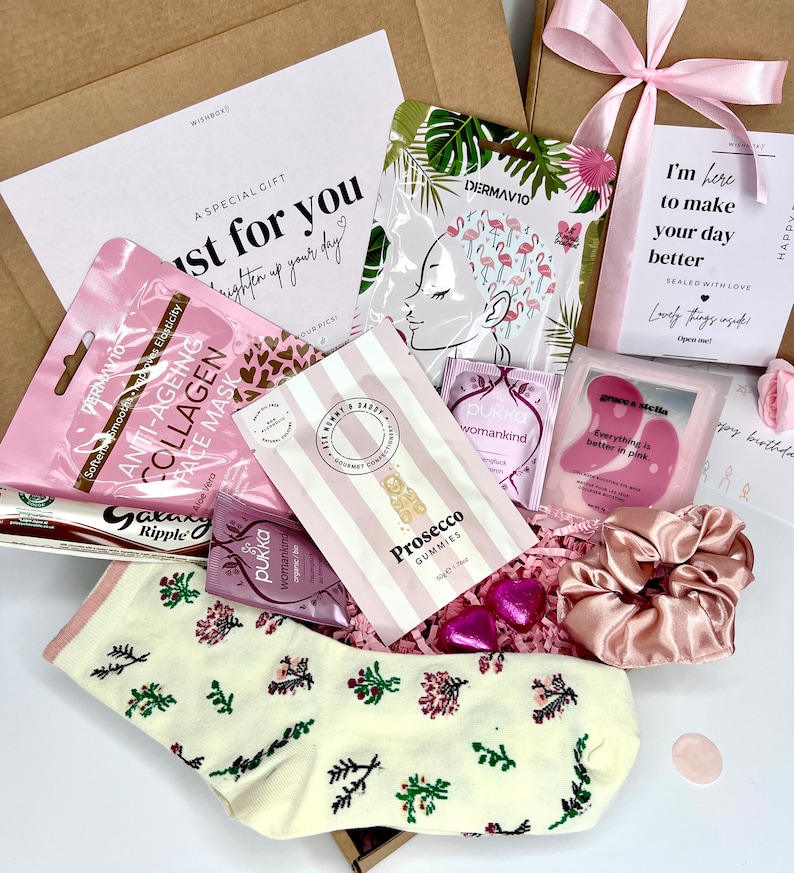 May include: A pink and white gift box filled with various items, including a face mask, a pink hair scrunchie, a bag of gummies, and a pair of socks with a floral pattern. The box is decorated with a pink ribbon and a card that says "I'm here to make your day better".