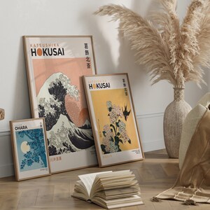 May include: Three framed prints of Japanese woodblock prints, featuring the Great Wave off Kanagawa, a hydrangea, and a moonlit scene. The prints are in a minimalist style with a peach, yellow, and blue colour scheme. The prints are hanging on a white wall with a vase of dried pampas grass in the foreground.