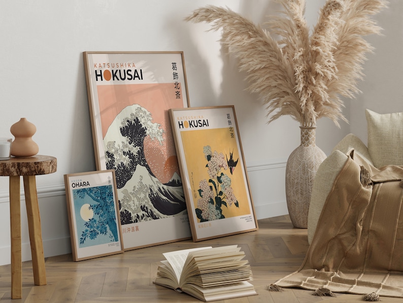 May include: Three framed prints of Japanese woodblock prints, featuring the Great Wave off Kanagawa, a hydrangea, and a moonlit scene. The prints are in a minimalist style with a peach, yellow, and blue colour scheme. The prints are hanging on a white wall with a vase of dried pampas grass in the foreground.
