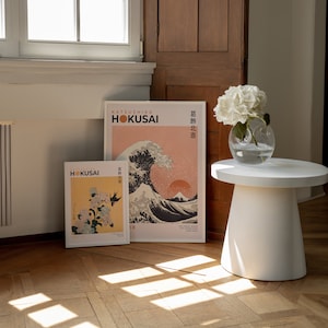 May include: Two framed prints of Japanese art by Katsushika Hokusai. The prints are on a light wood floor with a white side table and a vase of white flowers.