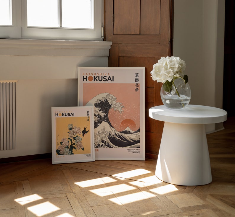 May include: Two framed prints of Japanese art by Katsushika Hokusai. The prints are on a light wood floor with a white side table and a vase of white flowers.