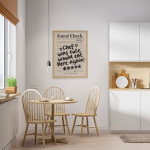 May include: A framed print with a white background and black text that reads "Guest Check" and "Chef was cute, would eat here again!" with five stars below the text. The print is hanging on a white wall in a kitchen setting with a wooden table and chairs.