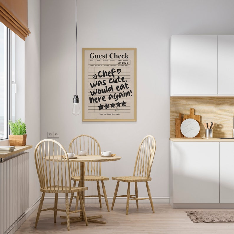 May include: A framed print with a white background and black text that reads "Guest Check" and "Chef was cute, would eat here again!" with five stars below the text. The print is hanging on a white wall in a kitchen setting with a wooden table and chairs.