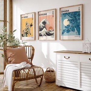 May include: Three framed prints of Japanese art hang on a white wall above a white cabinet with a wooden top. The prints feature a wave, a flowering tree, and a floral design. The prints are all in a similar style, with a white background and black outlines. The cabinet has two drawers and two doors.