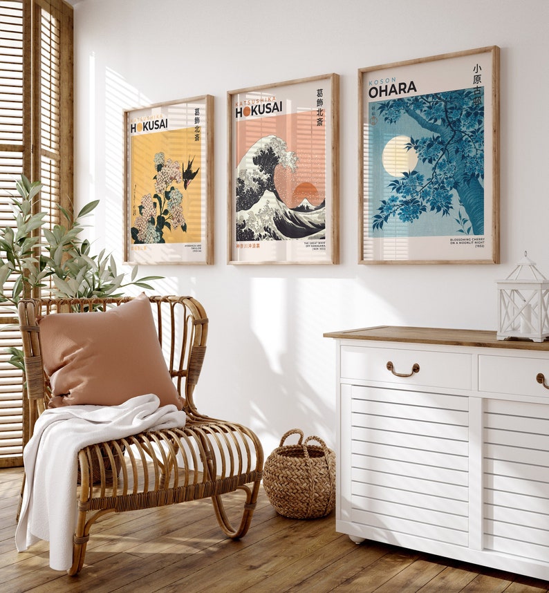 May include: Three framed prints of Japanese art hang on a white wall above a white cabinet with a wooden top. The prints feature a wave, a flowering tree, and a floral design. The prints are all in a similar style, with a white background and black outlines. The cabinet has two drawers and two doors.