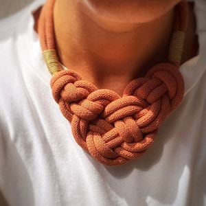 Può includere: Un primo piano di una collana di corda arancione annodata. La collana è realizzata in corda spessa intrecciata e ha un design semplice ed elegante.
