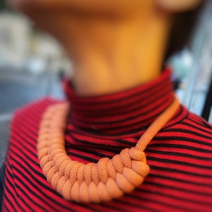 Può includere: Primo piano di una persona che indossa un maglione a collo alto a righe rosse e nere con una collana di corda spessa color pesca.