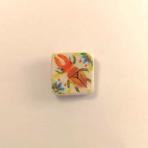 Può includere: Una piastrella quadrata in ceramica bianco sporco con un coleottero arancione dipinto a mano con grandi tenaglie e ali arancioni e verdi. La piastrella è decorata con piccoli fiori blu e punti gialli.