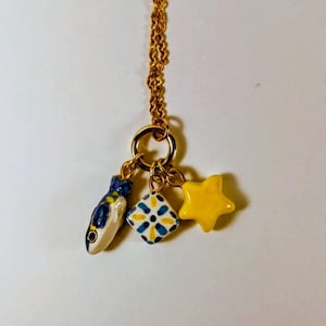 Può includere: Collana dorata con tre ciondoli in ceramica: un pesce blu e bianco, un quadrato con un motivo floreale blu e giallo e una stella gialla. I ciondoli sono sospesi a un anello e una catena dorati.