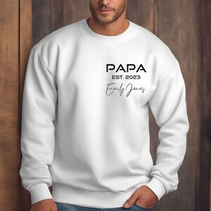 Könnte beinhalten: Weißes Sweatshirt mit dem schwarzen Aufdruck "PAPA EST. 2023 Emily Jones". Das Sweatshirt hat einen Rundhalsausschnitt und lange Ärmel mit gerippten Bündchen. Das Design ist mittig auf der Brust platziert.