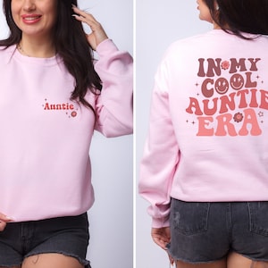 Könnte beinhalten: Ein hellrosa Sweatshirt mit dem Wort "Auntie" in roter Schrift auf der Vorderseite. Auf der Rückseite des Sweatshirts befindet sich der Schriftzug "In My Cool Auntie Era" in einem Retro-Schriftstil mit Blumen- und Smiley-Grafiken.