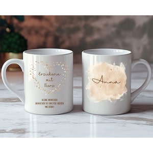 May include: Two white ceramic mugs with gold lettering. One mug says "Erzieherin mit Herz" and "Kleine Menschen brauchen so grosse Herzen wie deins!" The other mug says "Anna" in a gold script font.