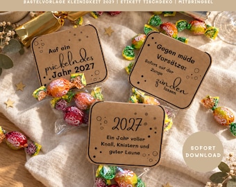 May include: Three square brown paper tags attached to clear plastic bags filled with colorful wrapped candies. The tags feature black printed text in German, including phrases like 'Auf ein prickelndes Jahr 2027' and 'Gegen müde Vorsätze'.