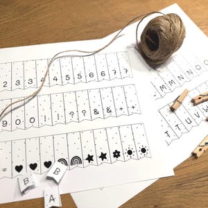 Puede incluir: Un papel blanco con n&uacute;meros, letras y s&iacute;mbolos impresos, incluyendo corazones, estrellas y arco&iacute;ris. Tambi&eacute;n hay una madeja de hilo y peque&ntilde;as pinzas de madera. El papel incluye las letras A, B y los n&uacute;meros 1-9.