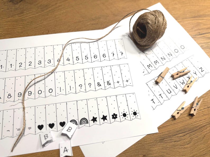 Puede incluir: Un papel blanco con n&uacute;meros, letras y s&iacute;mbolos impresos, incluyendo corazones, estrellas y arco&iacute;ris. Tambi&eacute;n hay una madeja de hilo y peque&ntilde;as pinzas de madera. El papel incluye las letras A, B y los n&uacute;meros 1-9.
