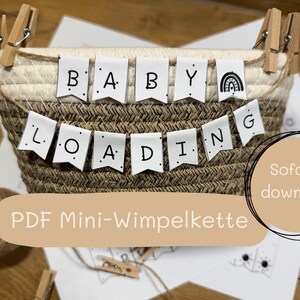 Puede incluir: Una cesta tejida con una pancarta que dice "BABY LOADING" en letras negras. La pancarta est&aacute; colgada en la parte superior de la cesta con peque&ntilde;as pinzas de madera. Tambi&eacute;n se ve una peque&ntilde;a bola de hilo y un papel con un dibujo.