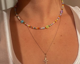 May include: A layered necklace with a beaded chain featuring colorful beads and smiley face charms. The bottom layer is a gold chain with a small cross pendant.