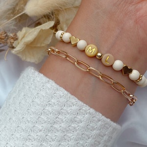 Könnte beinhalten: Ein weißes Perlenarmband mit goldenen Herzanhängern und goldenen Buchstabenperlen, die "M L L" buchstabieren. Das Armband wird an einem Handgelenk getragen, wobei ein goldenes Gliederarmband darüber gelegt wird.