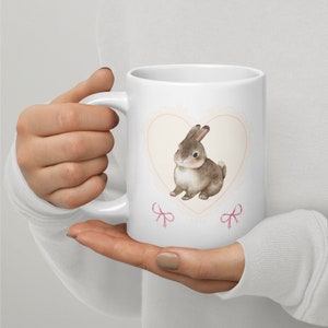 May include: White ceramic mug with a pink heart design featuring a brown bunny rabbit. The heart has a lace-like border and two pink bows.