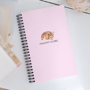 May include: A pink spiral-bound notebook with the text "I'M JUST A GIRL" and a watercolor illustration of a fawn. The notebook has a black spiral binding and is on a white surface.