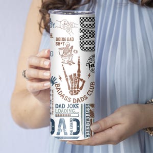 K&ouml;nnte beinhalten: Ein wei&szlig;er 590 ml Becher mit verschiedenen Grafiken und Texten, darunter "DAD", ein Skelett auf einer Toilette und der Ausdruck "BADASS DADS CLUB". Der Becher hat ein schwarz-wei&szlig;es Schachbrettmuster und eine braune Skelett-Handgeste.