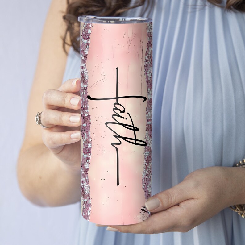 May include: A pink tumbler with the word "Faith" in black script. The tumbler has a glittery border and a light pink background. The tumbler is held by a person wearing a blue dress.