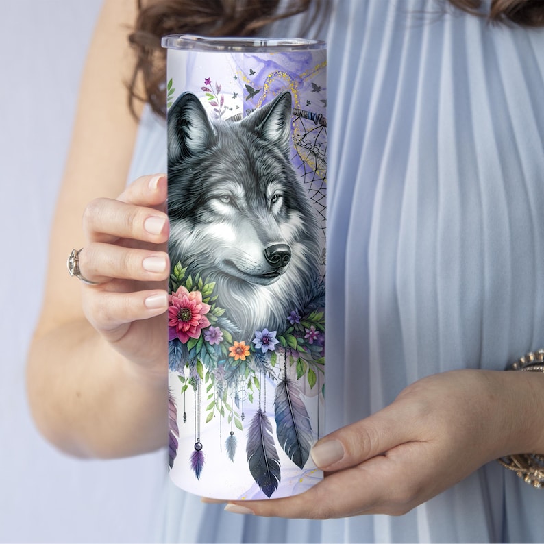 Puede incluir: Un vaso de acero inoxidable blanco con la cabeza de un lobo gris rodeada de flores y plumas de colores. Los ojos del lobo son azules y el fondo es de estilo acuarela morado y azul.