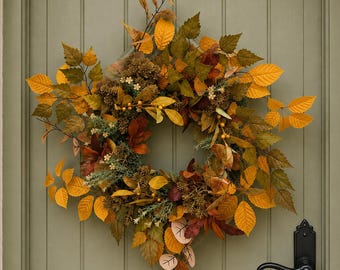 May include: A circular wreath composed of autumn-themed foliage, including serrated leaves in shades of yellow, orange, brown, and deep green. The arrangement features small clusters of berries and dried floral elements, creating a dense texture.