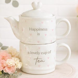 Puede incluir: Juego de té de cerámica blanca con tetera y taza apiladas. La tetera tiene una tapa en forma de corazón y corazones rosas. El texto "Happiness is" está en la tetera y "a lovely cup of Tea" en la taza. Sobre una base de madera.