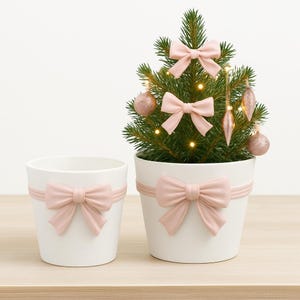 Può includere: Due vasi in ceramica bianca con fiocchi rosa. Uno contiene un piccolo albero di Natale decorato con fiocchi e ornamenti rosa. L'albero è illuminato da lucine, creando un'atmosfera festosa.