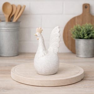 Könnte beinhalten: Eine weiße Keramik-Hahn-Figur mit detailliertem Federdesign, die auf einem runden Holzservierbrett steht. Der Hahn hat einen cremefarbenen Kamm und Schnabel. Der Hintergrund umfasst ein Holzschneidebrett und einen Metalleimer mit Holzlöffeln.