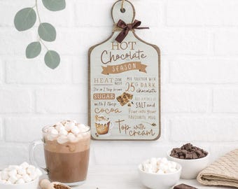 May include: A wooden sign with a brown ribbon and rope for hanging, featuring the text "HOT Chocolate SEASON" and a recipe. A mug of hot chocolate with marshmallows, bowls of marshmallows and chocolate, and a wooden scoop are on a white surface.