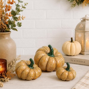 May include: Three ceramic pumpkins with glossy tan bodies and dark green stems. The pumpkins vary in size, featuring deep vertical ridges and a smooth, reflective finish. They are arranged in a cluster, showcasing their rounded shapes and distinct, realistic stem details on top of each piece.