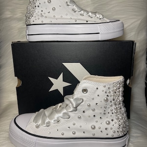 May include: White high-top sneakers with a platform sole, embellished with pearls and rhinestones. The sneakers have a satin ribbon tied around the laces.