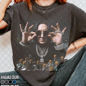 Può includere: T-shirt grigio scuro con un ritratto di un uomo con occhiali da sole e gioielli, con diverse piccole bottiglie davanti. La maglietta ha il testo "AVAILABLE COLORS" e "PEPPER COLORS" con campioni di colore.