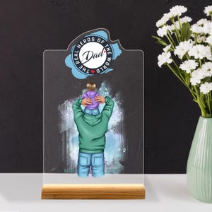 Könnte beinhalten: Eine klare Acrylplatte mit Holzsockel zeigt eine farbenfrohe Illustration eines Vaters, der sein Kind auf den Schultern trägt. Die Platte hat oben einen Kreis mit dem Text "The Real Heroes of the World Dad" in Schwarzweiß mit einem roten Herzen.