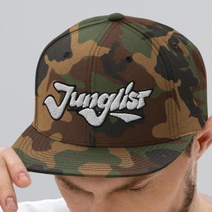 Op de afbeelding: Een baseballcap met camouflage-print en een wit geborduurd logo met de tekst "Junglist".
